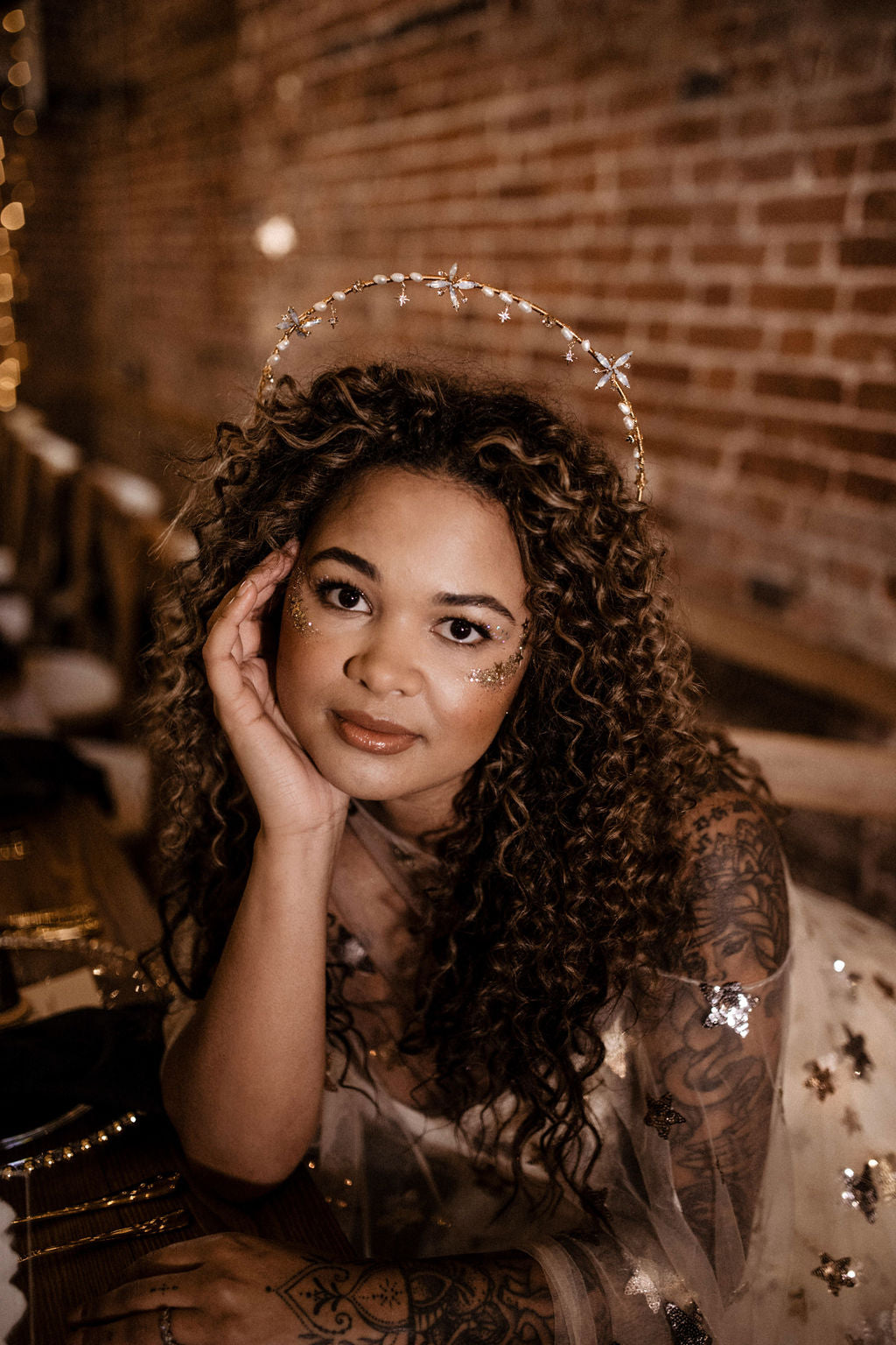 A woman with curly brown hair wearing a silver tiara and sparkly embellished dress, posing in front of a warm brick wall background.