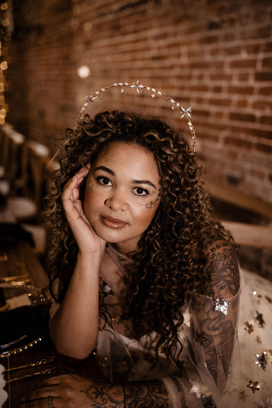 A woman with curly brown hair wearing a silver tiara and sparkly embellished dress, posing in front of a warm brick wall background.